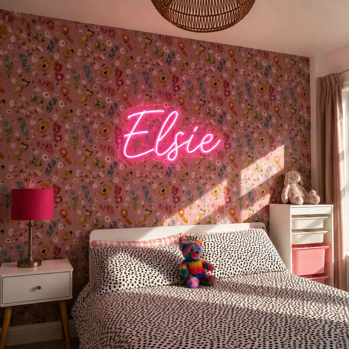 A pink neon sign reading 'Elsie' mounted on a pink floral wallpapered wall in a styled bedroom. The room features a bed with black-and-white patterned bedding, a white nightstand with a pink lamp, and a small stuffed animal on the bed.