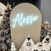 A festive holiday backdrop setup featuring a large beige arched panel with a glowing light blue script neon sign reading 'Alesso', flanked by decorated flocked Christmas trees and small white holiday houses.