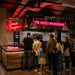 A group of people standing in a restaurant or cafe queue area, facing a counter with neon signs including 'Smash Burgers', 'MR HUGO SMOKEHOUSE', and 'Milkshakes'. White dimension lines with text labels '80cm wide', '160cm wide', and '60cm wide' are overlaid on the scene.