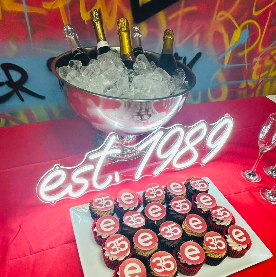 A celebratory tabletop setup with a large silver ice bucket holding champagne bottles, a red tablecloth, a white plate of cupcakes topped with red circles and white script reading '35', and a glowing neon-style 'est. 1989' sign in front.