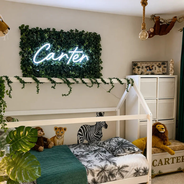 A children's bedroom featuring a white house-frame bed with tropical-print bedding, surrounded by plush animal toys. Above the bed is a green faux foliage wall neon sign with the name 'Carter' in glowing white script.
