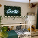 A children's bedroom featuring a white house-frame bed with tropical-print bedding, surrounded by plush animal toys. Above the bed is a green faux foliage wall neon sign with the name 'Carter' in glowing white script.