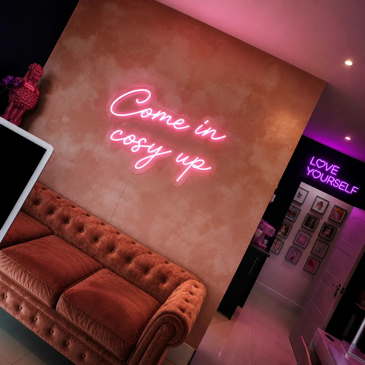 A pink neon sign reading 'Come in cosy up' mounted on a textured terracotta wall above a tufted brown sofa in a modern room with purple ambient lighting.