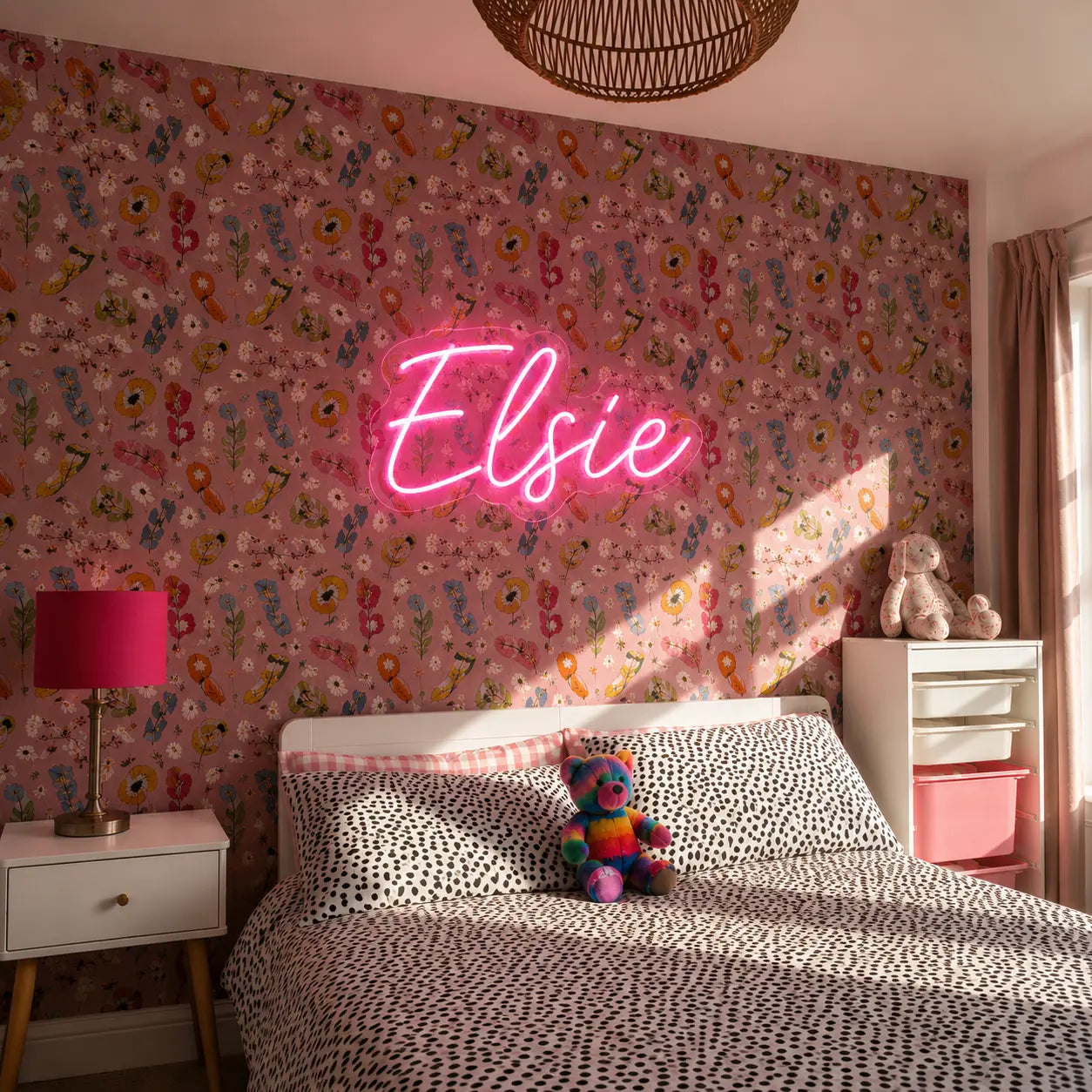 A pink neon sign reading 'Elsie' mounted on a pink floral wallpapered wall in a styled bedroom. The room features a bed with black-and-white patterned bedding, a white nightstand with a pink lamp, and a small stuffed animal on the bed.