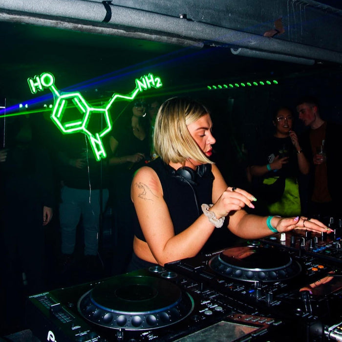 A woman DJing in front of a green Serotonin Neon Sign by clubserotone