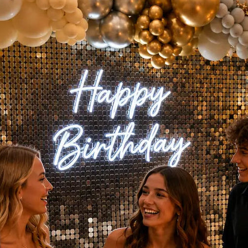 A group of friends celebrating a 16th birthday at an outdoor patio, gathered around a glowing 'Happy Birthday' sign. The background features a large balloon garland in gold, white, and silver, a large illuminated '16' sign, and a fire pit table with drinks.