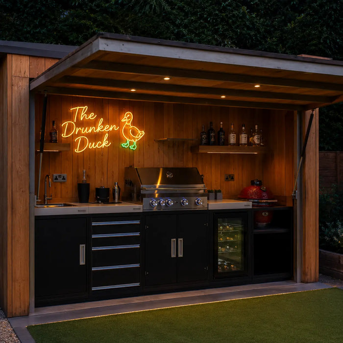 Outdoor kitchen bar setup with wooden cabinetry, stainless steel grill, sink, shelves, and a glowing neon sign reading “The Drunken Duck” with a duck neon sign. The scene is set at night in a backyard with artificial turf and dark foliage in the background.