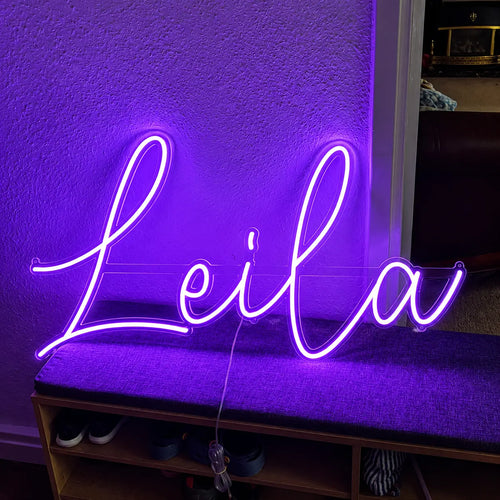 A purple neon sign spelling the name 'Leila' in cursive script, illuminated against a textured wall above a bench.