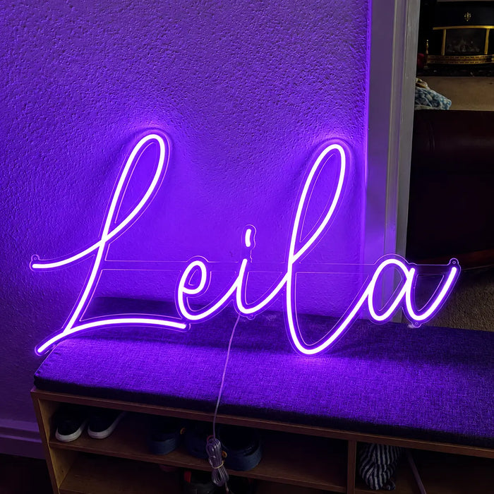 A purple neon sign spelling the name 'Leila' in cursive script, illuminated against a textured wall above a bench.