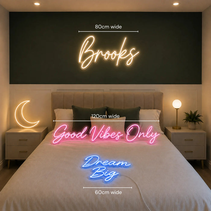 A bedroom scene showing a large glowing neon-style wall sign above the bed with the word 'Brooks' and dimension lines reading 80cm wide. A crescent moon lamp glows on the left nightstand, and a pink neon sign across the bed reads 'Good Vibes Only' with a 120cm width measurement.