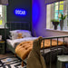 A styled children's bedroom featuring a metal twin bed with white bedding and a mustard yellow tufted comforter. A glowing blue neon sign reading 'OSCAR' is mounted on the dark wall above the bed.