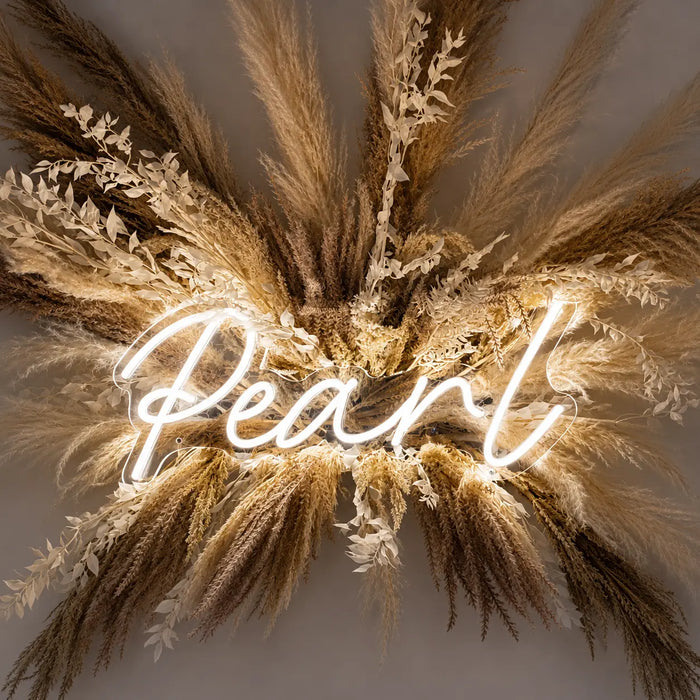 A decorative wall sign featuring the word 'Pearl' in glowing white neon script, surrounded by dried pampas grass and small white floral accents against a warm neutral background.