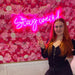 Woman posing in front of a pink flower wall with a pink Stay Weird Neon Sign by cakesadaisy