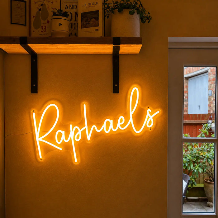 A glowing yellow-orange neon-style wall sign spelling "Raphaels" mounted on a warm beige wall, with a wooden shelf and framed decor above and a window showing an outdoor patio on the right.