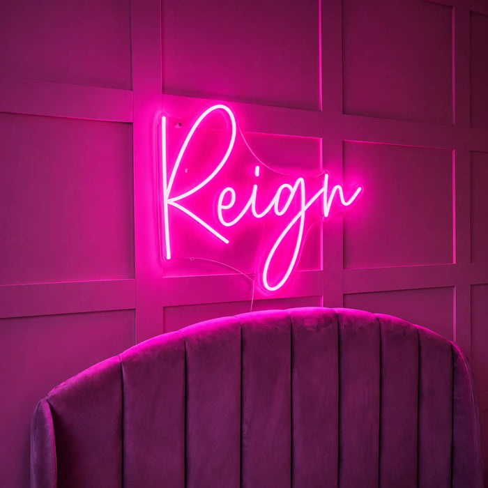 A bright pink neon sign spelling the word 'Reign' in cursive script, mounted on a dark paneled wall above a purple velvet upholstered chair.