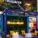 A booth display at an exhibition with a blue-framed wall covered in greenery and illuminated neon signs reading phrases such as 'This is not a sales pitch,' 'Hello!,' and 'Lounge.' Yellow chairs and white tables are arranged inside, with size labels overlaid: 200cm wide, 60cm wide, and 80cm wide.