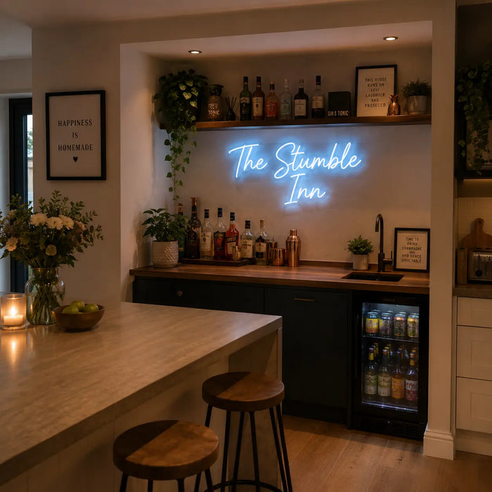 A glowing blue neon sign reading 'The Stumble Inn' mounted on a back wall above a home bar setup with bottles, plants, shelves, and stools in a warm, modern interior.