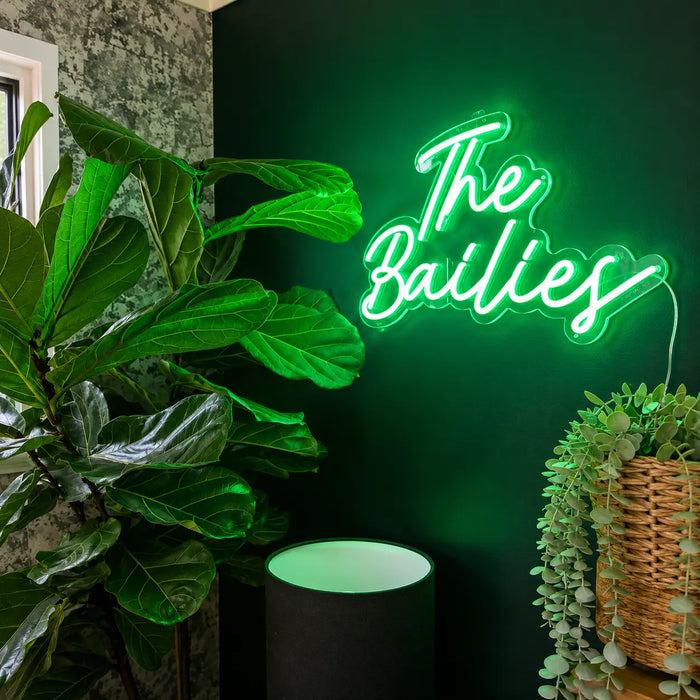 A glowing green neon-style wall neon sign reading “The Bailies” mounted on a dark green wall in a styled interior with large green plants and a wicker basket planter.