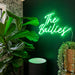 A glowing green neon-style wall neon sign reading “The Bailies” mounted on a dark green wall in a styled interior with large green plants and a wicker basket planter.