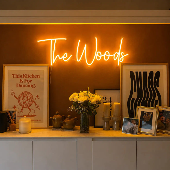 A glowing orange neon sign reading 'The Woods' mounted on a dark brown wall above a console table. The table is styled with framed art, candles, a vase of yellow flowers, and small decorative objects in a warm, cozy interior setting.