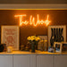 A glowing orange neon sign reading 'The Woods' mounted on a dark brown wall above a console table. The table is styled with framed art, candles, a vase of yellow flowers, and small decorative objects in a warm, cozy interior setting.