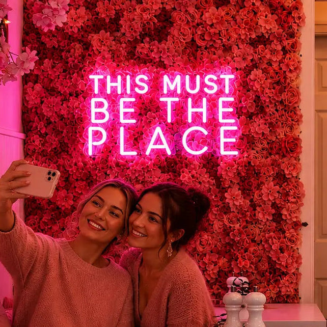 Two women taking a selfie in a cozy, pink-lit cafe or restaurant corner with a floral wall, a pink neon sign reading 'THIS MUST BE THE PLACE,' and tables with cups and white salt and pepper shakers in the foreground.