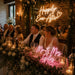 A wedding reception scene with a bride and groom seated at a decorated head table, surrounded by guests in a warmly lit brick-walled hall. Neon signs on the table read 'Happily Ever After' and 'Mr.