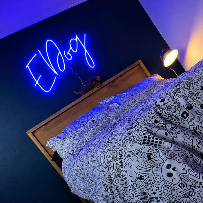 A blue neon sign spelling 'EDog' mounted on a dark wall above a bed with a patterned duvet and wooden headboard, with a bedside lamp glowing nearby.