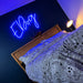 A blue neon sign spelling 'EDog' mounted on a dark wall above a bed with a patterned duvet and wooden headboard, with a bedside lamp glowing nearby.