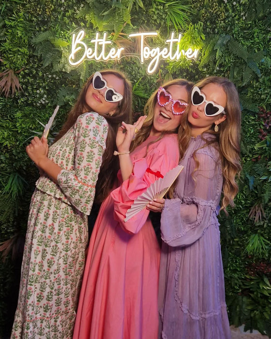 Three women posing against a plant backdrop with a custom neon sign saying "Better Together"