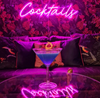 Pink "cocktails" neon sign against bold leaf wallpaper and sofa. Blue cocktail with orange curl in foreground.