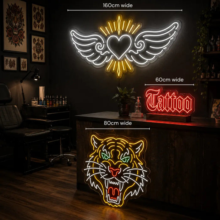 Three neon wall signs displayed in a tattoo studio setting: a large winged heart sign reading 160cm wide, a red 'Tattoo' script sign reading 60cm wide, and a tiger head sign reading 80cm wide. The studio features a dark wood counter, tattoo chair, and framed artwork on the wall.