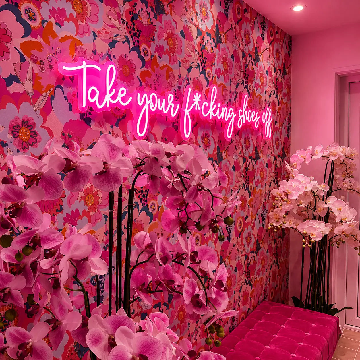 A vibrant pink neon sign reading 'Take your shoes off' mounted on a wall covered in dense pink floral wallpaper. Large pink artificial orchids are arranged in the foreground, with a bright pink tufted bench in the lower right corner.