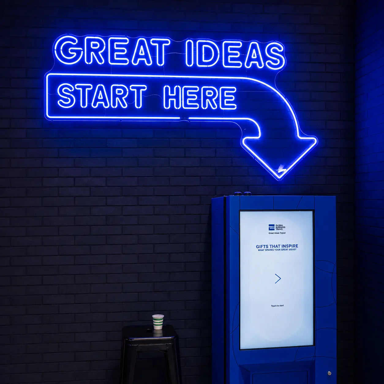 A blue neon sign on a dark brick wall reads 'GREAT IDEAS START HERE' with an arrow pointing down toward a blue kiosk or touchscreen display.