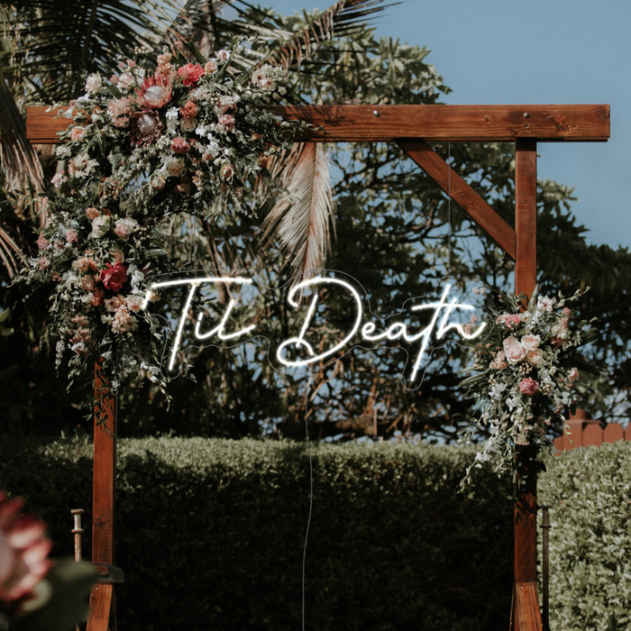 Til Death LED neon sign in snow white hung from a wooden frame with flowers against a tropical background