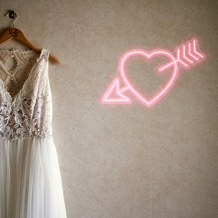Pastel pink neon sign heart with arrow hanging on plain wall next to a white wedding dress