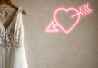 Pastel pink neon sign heart with arrow hanging on plain wall next to a white wedding dress